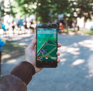 A person is holding a smartphone with a gaming app open, featuring an augmented reality map. The setting is outdoors, with people visible in the background, suggesting a park or public space.