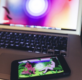 A smartphone displaying an app or game with cartoonish characters and greenery is resting on a laptop keyboard. The laptop screen shows a blurred, colorful background with a circular design.