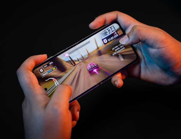 Hands holding a smartphone with a racing game displayed on the screen. The image shows a vibrant racing scene with a red and white car moving at high speed along a desert-like track. Various on-screen controls and speed indicators are visible.